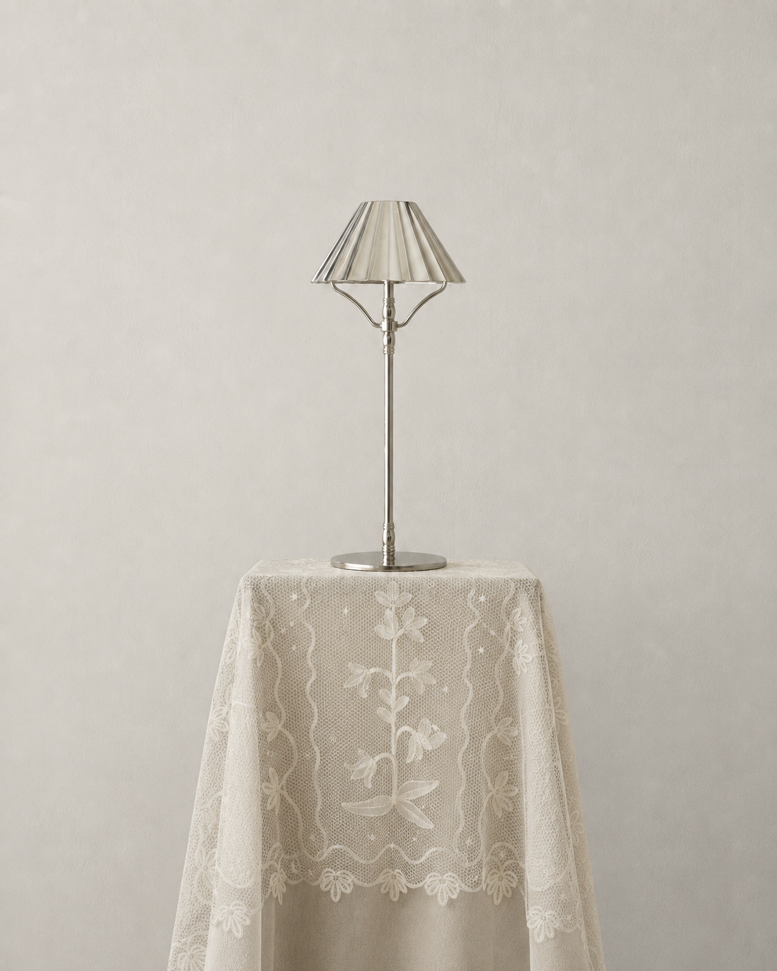 A silver cordless table lamp with a pleated shade and round base sits on top of a draped cream lace tablecloth against a light gray wall.
