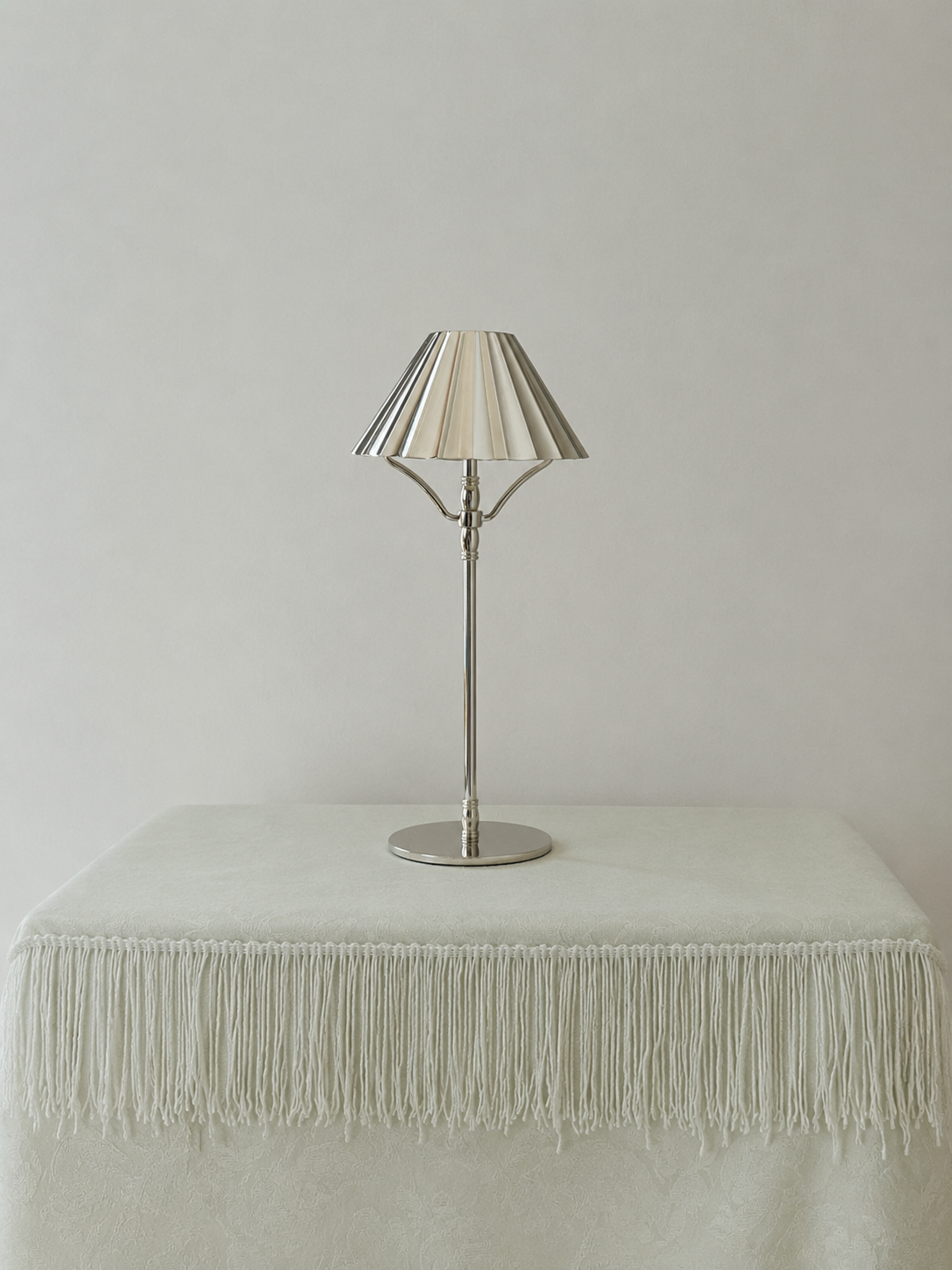 A modern cordless silver table lamp with a pleated fabric shade in a warm cream tone and a polished silver metal stand, displayed on a cream fringed tablecloth against a neutral light gray background.