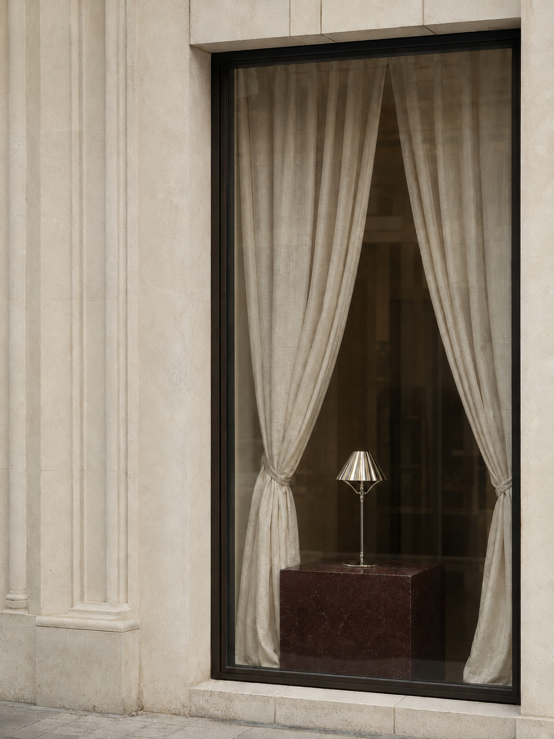 A tall window with beige curtains tied back, showing a small cordless silver table lamp on a dark marble pedestal inside. The window is set into a light stone building facade.
