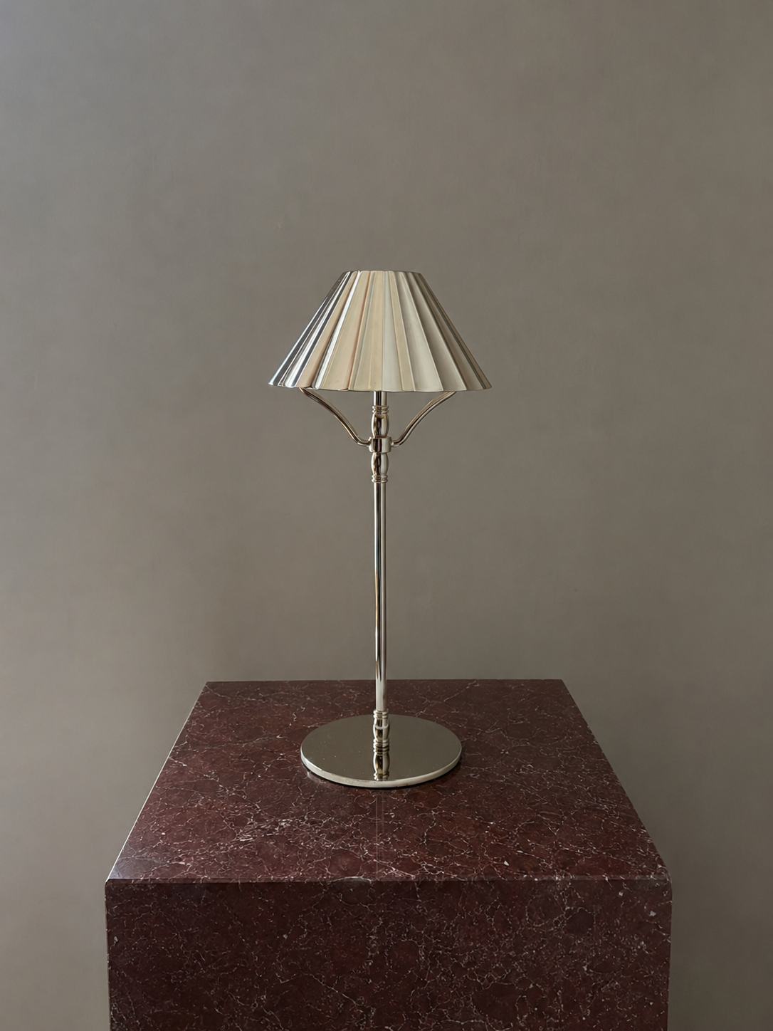 A modern silver cordless table lamp with a pleated cream fabric shade and a polished silver metal stand, displayed on a dark red marble pedestal against a neutral gray background.