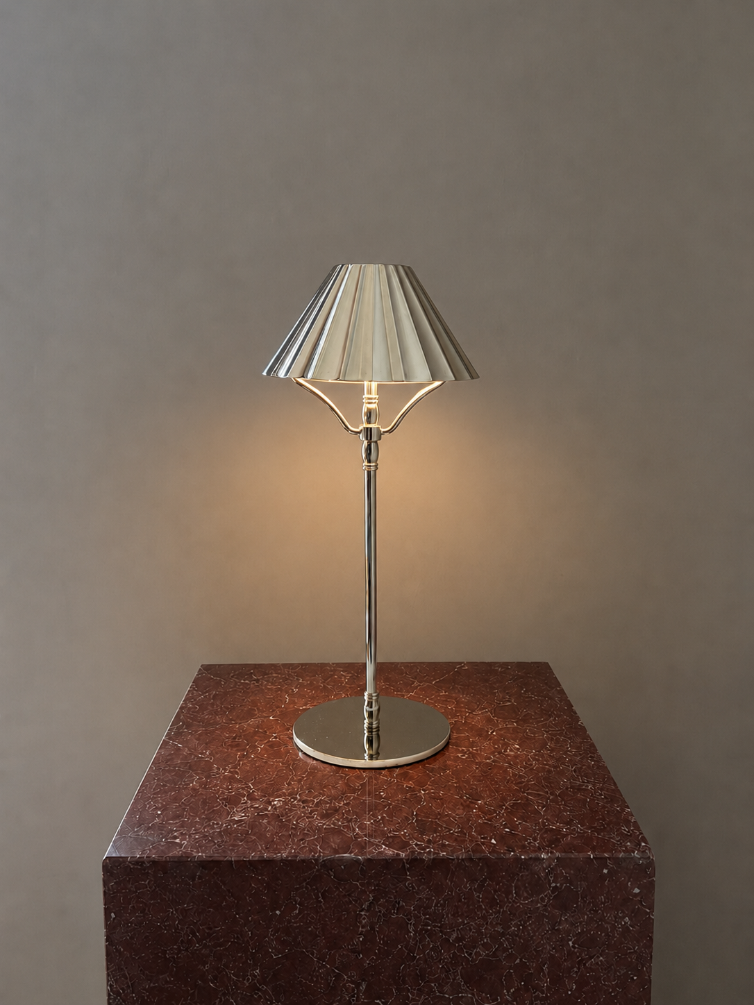 A modern cordless table lamp with a pleated metallic silver shade and a polished brass stand, illuminated with a warm glow, displayed on a dark reddish-brown marble pedestal against a neutral gray background.